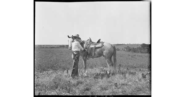 A black-and-white image of a cowboy standing in a field next to his horse adjusting the saddle.