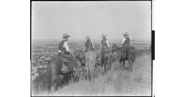 A black-and-white photograph of four cowboys on horseback, viewed from behind, looking over plains in the distance.