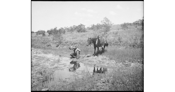 A black-and-white image of a cowboy crouched next to a stream drinking water from his hat as his horse stands behind him.