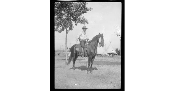 A black-and-white image of a man wearing a cowboy hat seated on a horse; several tipis in the background.