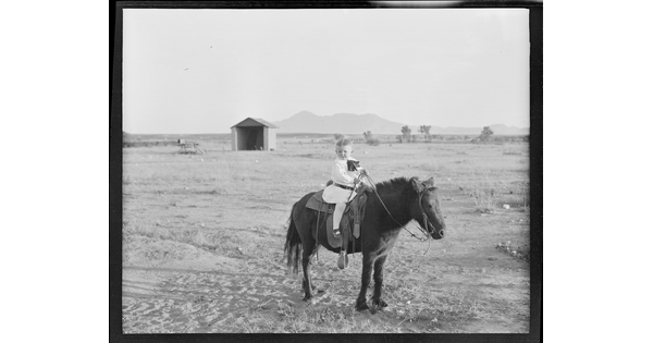 A black-and-white image of a young White child on a pony in a dusty field.