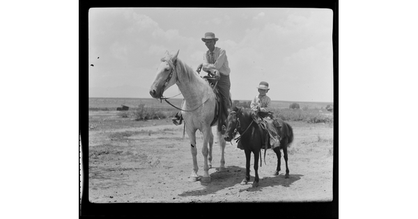 A black-and-white image of a White man wearing a cowboy hat on horseback and, next to him, a White child wearing a cowboy hat on a pony.