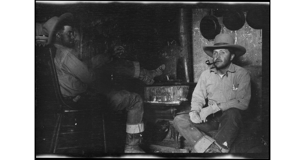 A black-and-white image of two White cowboys seated inside: one on a chair with his foot propped on a stove, the other next to the stove smoking a pipe.