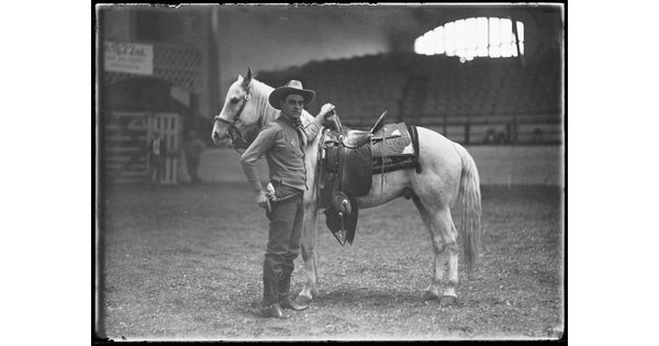 A black-and-white image of a White cowboy standing next to his saddled horse in an empty arena.