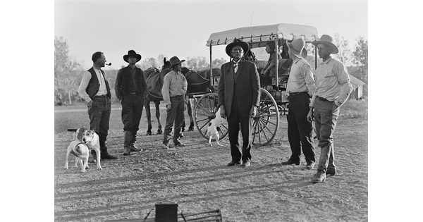 A black-and-white image of six men dressed in western attire and several dogs standing in front of a carriage.