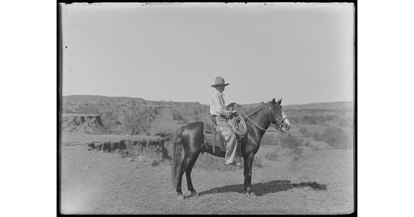 A black-and-white image of a cowboy on horseback standing, in profile, on a plain.