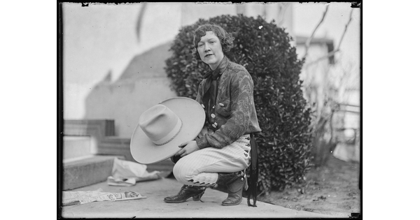 A black-and-white image of a White woman wearing cowboy boots and holding a large cowboy hat as she squats in front of a shrub on a sidewalk.