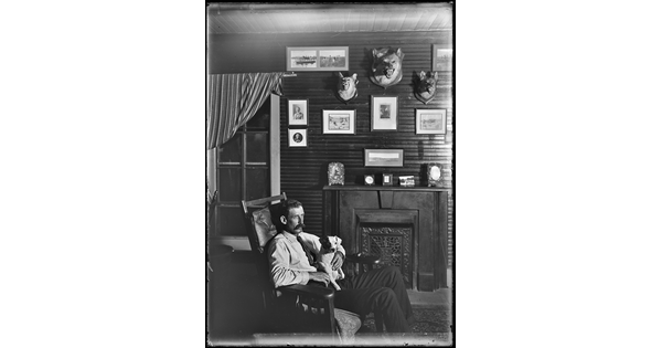 A black-and-white image of a White man seated in a leather chair with a small dog on his lap; in the background is a fireplace beneath framed art and mounted animal heads.