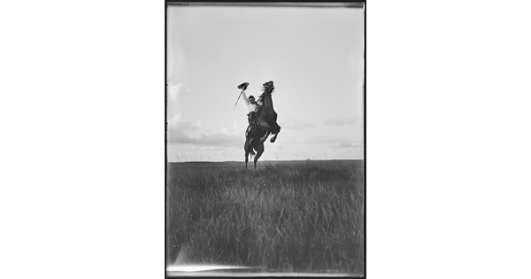 A black-and-white image of a White man on a rearing horse holding his hat in one hand above his head.
