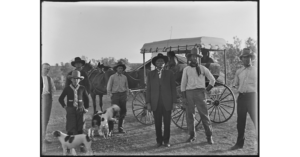 A black-and-white image of six men dressed in western attire and several dogs standing in front of a carriage.