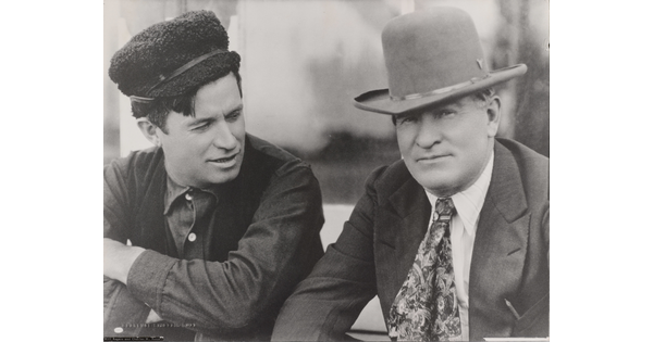 A black-and-white photograph of two White men: the man on the left wearing a fuzzy hat, dark vest, and button-down shirt; the man on the right wearing a cowboy hat with a jacket and tie.