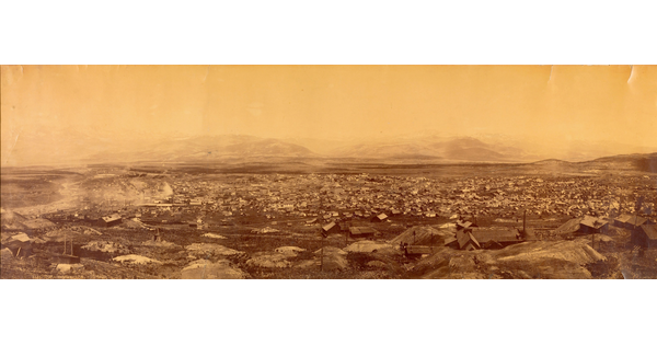 A sepia-toned panoramic photograph taken from high up of a small town in a barren, rocky landscape with mountains in the distance.