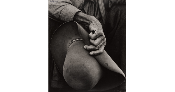 A close-up black-and-white photograph of a hand with aged, wrinkled skin holding a felt cowboy hat.