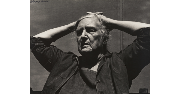 A black-and-white photograph of an older White woman, tired expression on her face, wearing a denim jacket, her hands folded on top of her head.