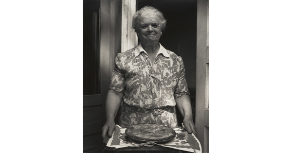 A black-and-white photograph of a smiling older White woman wearing a floral print dress and holding out a tray with a pie on it.