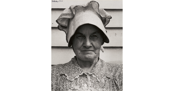 A black-and-white photograph of an older White woman, head and shoulders filling the frame, wearing a bonnet and a printed blouse.
