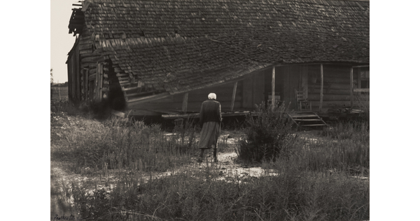 A black-and-white photograph of a hunched figure walking toward a log cabin with a collapsed roof and a rocking chair on the porch.