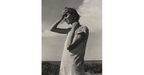 A black-and-white photograph of a White woman standing outside, holding the side of her neck with one hand and her forehead with her other.