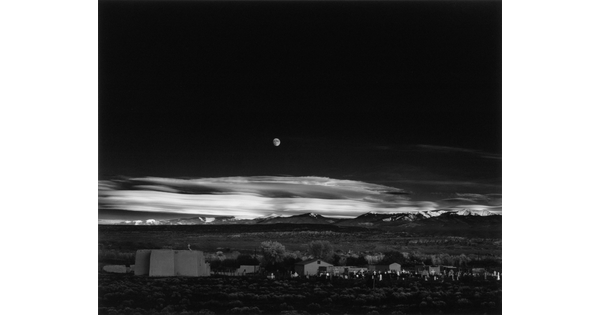 A black-and-white landscape photograph of a town with mountains in the background and the moon rising above the clouds in the night sky.
