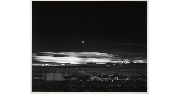 A black-and-white landscape photograph of a town with mountains in the background and the moon rising above the clouds in the night sky.