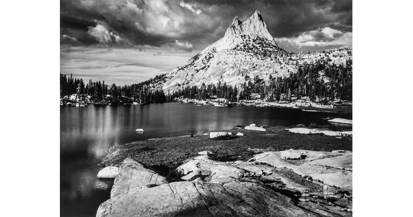 A black-and-white photograph of a rocky mountain peak from across a still lake surrounded by trees.