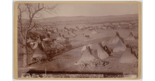 A sepia-toned photograph of many tipis and wagons on a plain.