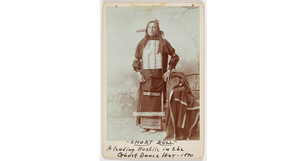 A sepia-toned full-body portrait of an Indigenous man in cultural dress, including a breastplate.