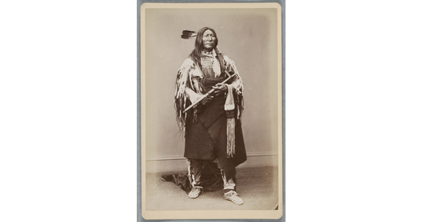 A sepia-toned full-body portrait of an Indigenous man in cultural dress holding a pipe.