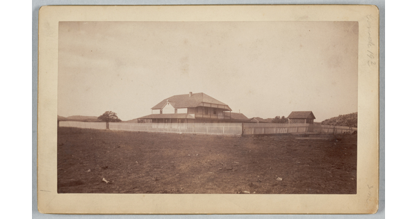 A sepia-toned photograph of a two-story house behind a picket fence on an open field.