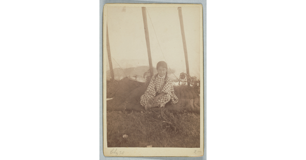 A sepia-toned photograph of a young girl wearing a gingham dress and sitting cross-legged on a blanket.
