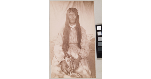 A sepia-toned portrait of a seated Indigenous woman with long dark hair wearing a long-sleeved gingham dress.
