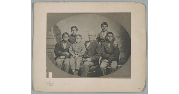 A black-and-white studio portrait photograph of a seated White man surrounded by five Indigenous boys of various ages.