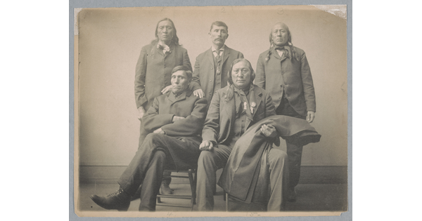 A sepia-toned studio portrait photograph of five Indigenous men in dark suits, three standing and two seated.