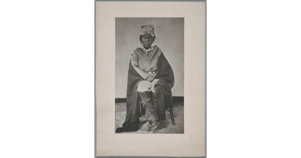 A black-and-white portrait photograph of a seated Native American man wearing a head covering and with a blanket wrapped around is shoulders.
