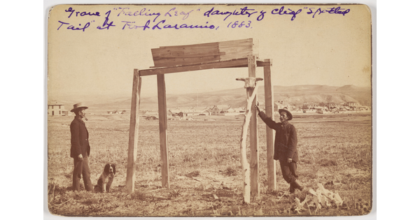 A sepia-toned photograph of two men and a dog next to a simple wood structure on the outskirts of a town.