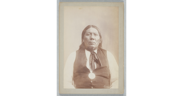 A black-and-white portrait photograph of an Indigenous person with long hair and earrings wearing a white shirt under a buttoned vest.