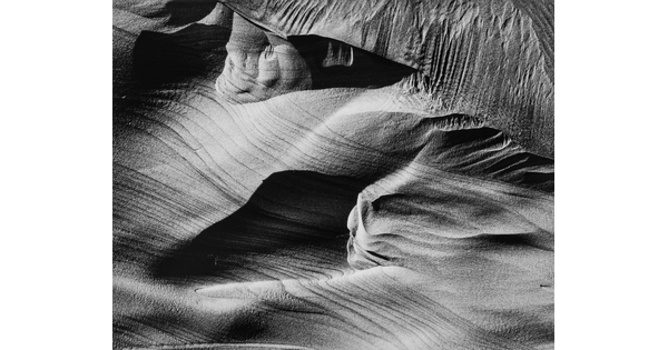 An abstract black-and-white photograph of sand dunes with horizontal and vertical striations that make it seem to be in motion.