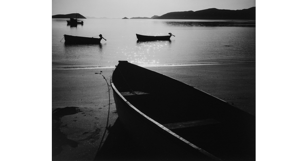 A black-and-white photograph of a rowboat on a sandy beach and three more out on still water with hills in the distance.