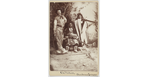 A sepia-toned photograph of three people, one dressed in western attire with a pistol at his side, and two in Indigenous people in cultural dress holding rifles.
