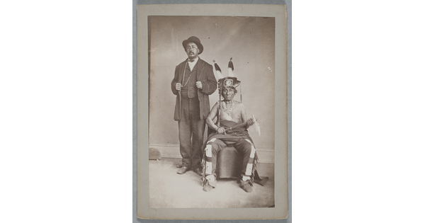 A black-and-white photograph of two Indigenous men: the one standing is wearing a bowler hat and a three-piece suit; the one seated wears a feathered hat, many necklaces, fringed pants, and holds a tomahawk in his lap.