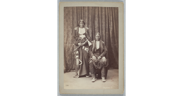A sepia-toned portrait photograph of two Indigenous people: one seated with a hat on their knee, and the other standing holding a tomahawk.