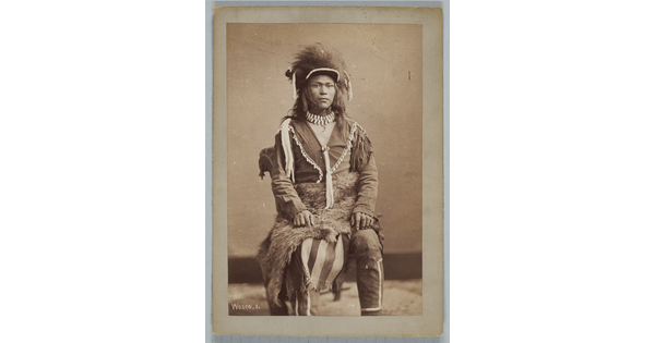A sepia-toned portrait photograph of a young, Indigenous man seated, wearing traditional dress.