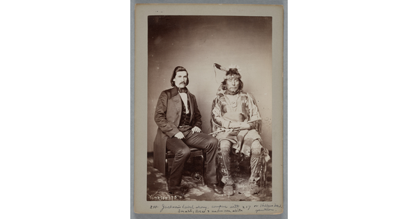 A sepia-toned portrait photograph of two seated men: a White man wearing a long suit jacket over a vest, and the other an Indigenous man in traditional dress holding a pipe.