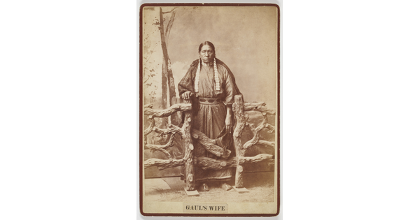 A sepia-toned studio photograph of an older Indigenous woman wearing a long polka-dotted dress, blanket around her shoulders, standing behind a wood fence.