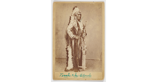 A sepia-toned portrait photograph of an Indigenous chief standing in traditional regalia, including a full-feathered headdress.