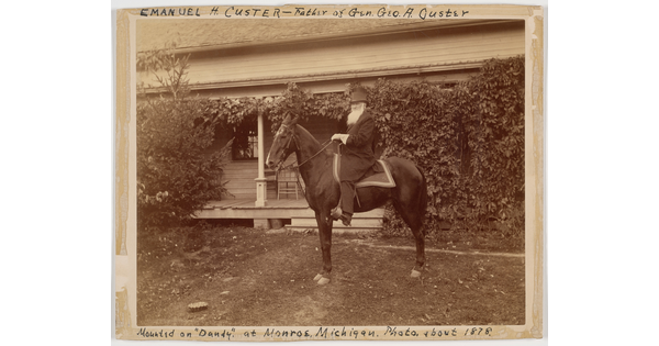A sepia-toned photograph of a bearded White man seated on a horse in front of a house covered in vines.
