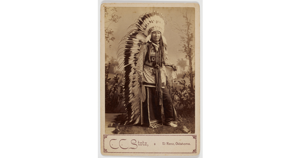 A sepia-toned studio portrait photograph of an Indigenous man in full traditional regalia, including a long, feathered headdress.