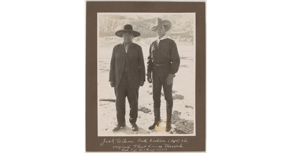 A black-and-white photograph of an Indigenous man in a suit and wide-brimmed hat standing next to a White man in dark clothes and a cowboy hat.