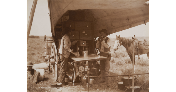 A sepia-toned photograph of two men eating and drinking from a chuckwagon as a horse stands in the background.