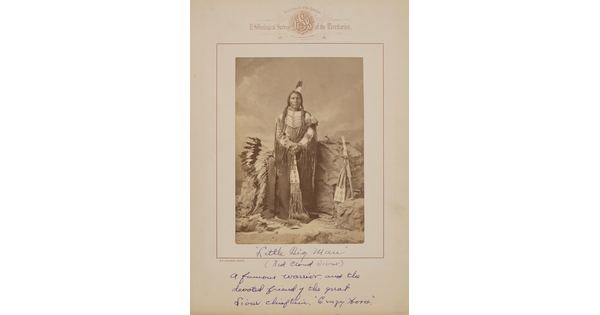 A black-and-white formal portrait photograph of an Indigenous man in traditional clothing surrounded by a headdress and several Indigenous objects.